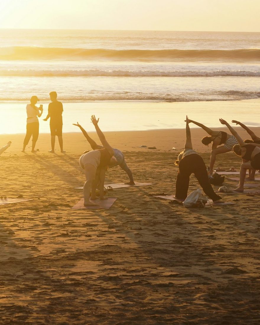 A small group doing yoga on the sand as the sun sets over the sea